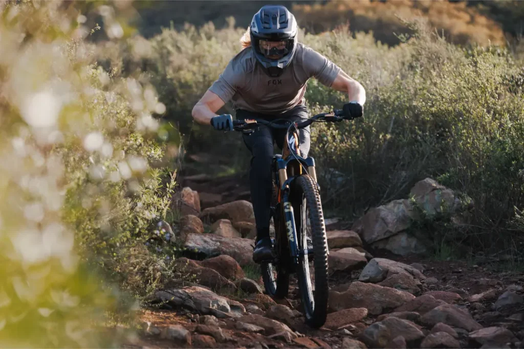Persona con casco y guantes conduce una bicicleta de montaña Enduro con horquilla FOX 38 2027, manteniendo el control en un sendero rocoso rodeado de arbustos.