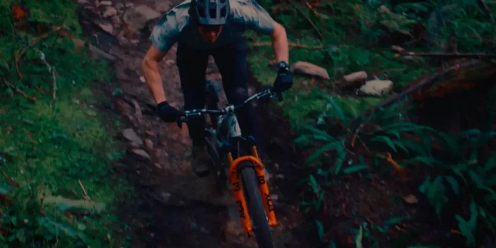 Una persona con casco y guantes conduce una bicicleta de montaña Enduro con un control preciso por un sendero rocoso y boscoso, equipada con la avanzada horquilla FOX 38 2027.