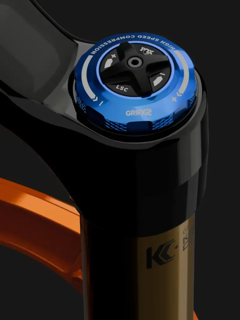 Primer plano de un mando de ajuste de horquilla de suspensión FOX 38 2027 con dial azul etiquetado "High Speed Compression" y "GRIPX2", que ofrece un control preciso de enduro en una horquilla de bicicleta negra y naranja.