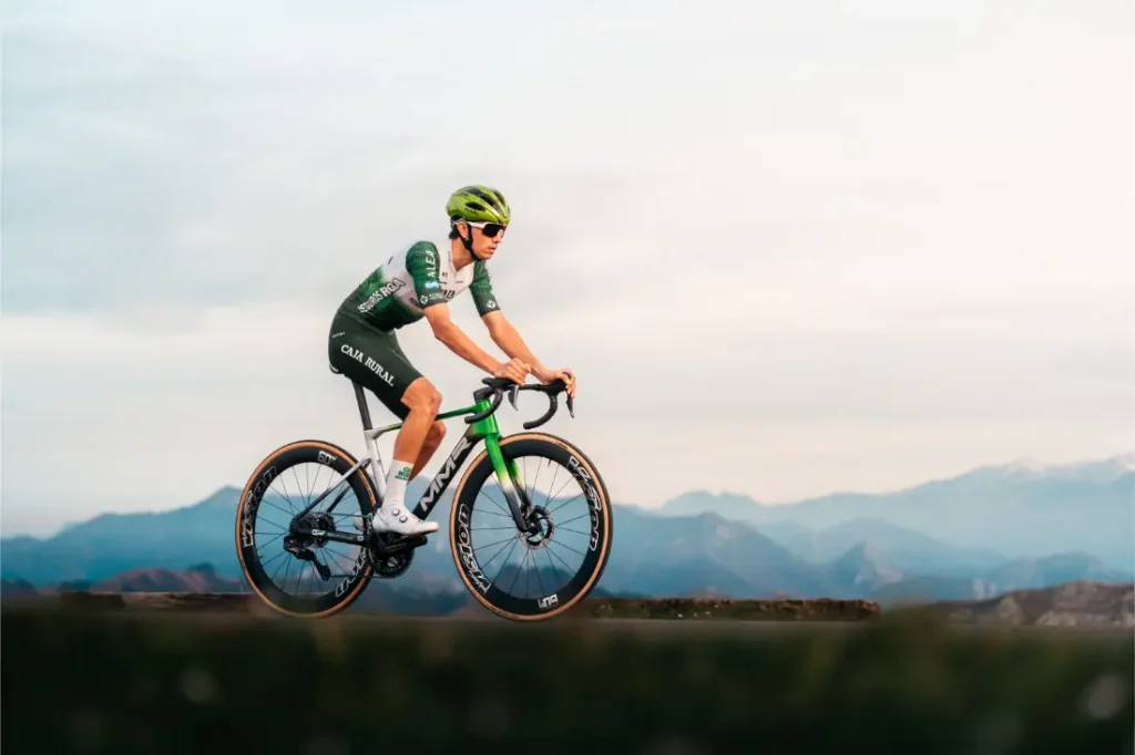 Un ciclista vestido de verde y blanco conduce una bicicleta de carretera MMR Aelion SL por un sendero con montañas y un cielo nublado de fondo.