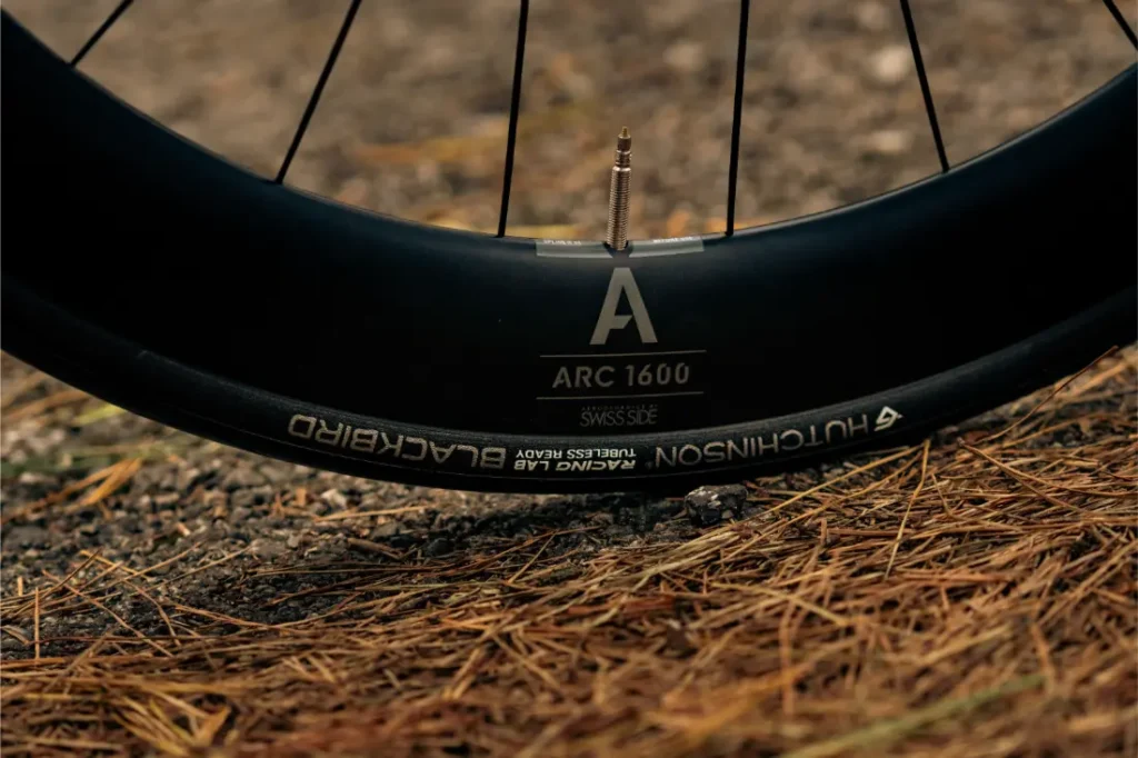 Primer plano de una rueda de bicicleta que muestra el vástago de la válvula, la llanta negra con la etiqueta "ARC 1600" y el neumático con la marca "Hutchinson Blackbird", con agujas de pino y gravilla en el suelo: perfecta para tu próximo paseo con la MMR Aelion SL de bici total.