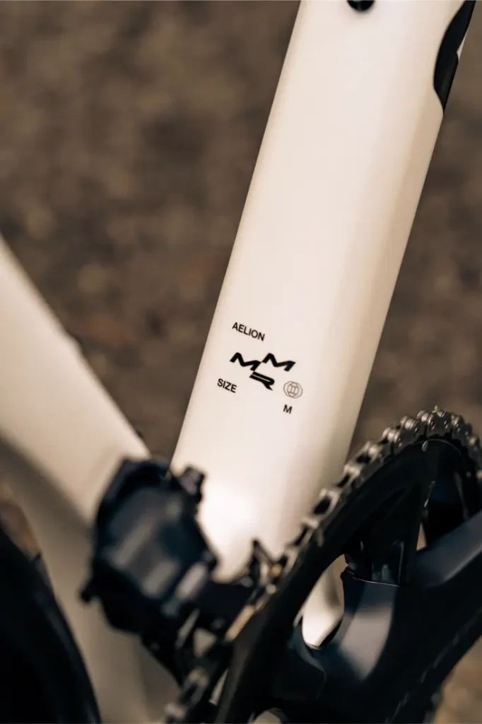 Primer plano de un cuadro de bicicleta MMR Aelion SL blanco cerca de la biela, con la marca "AELION", un gráfico y la etiqueta de la talla "M". Perfecta para quienes buscan un rendimiento total en bicicleta.