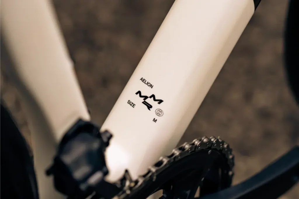 Primer plano de un cuadro de bicicleta en el que aparece la etiqueta "AELION SIZE M" junto a una cadena y un pedal, lo que pone de relieve el tamaño mediano de esta bici total MMR Aelion.