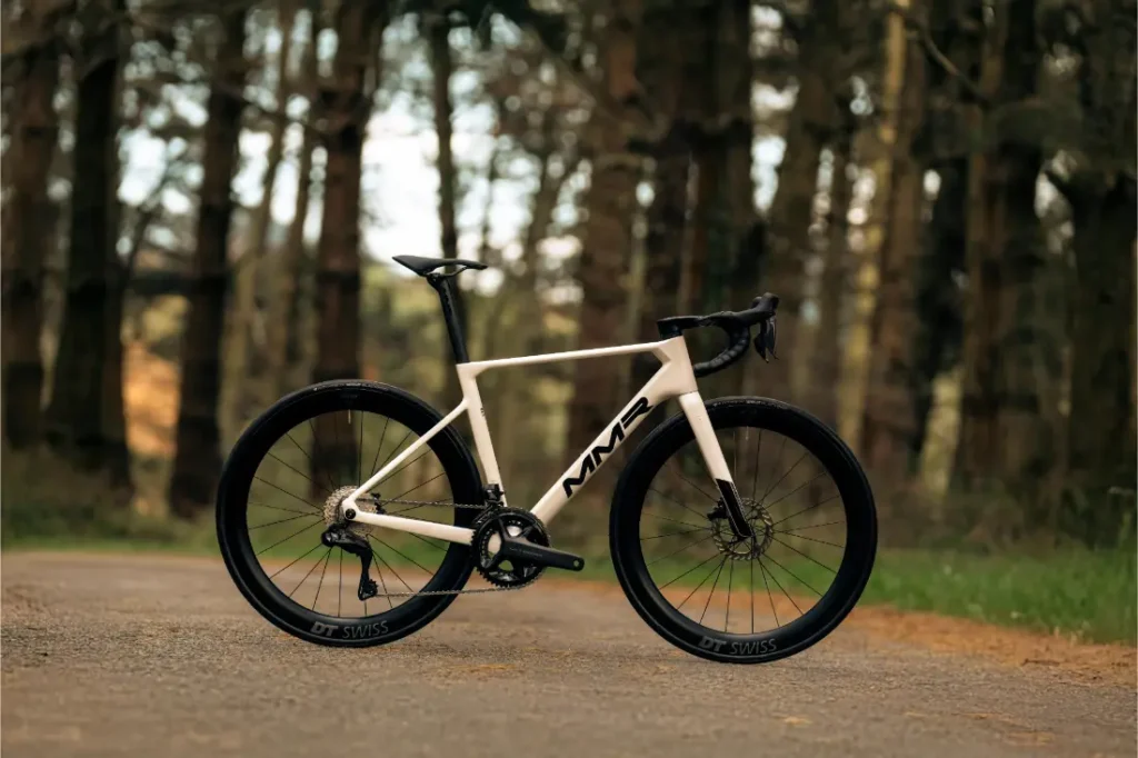 Una bicicleta de carretera MMR Aelion blanca con ruedas negras y manillar caído está aparcada en un camino pavimentado en una zona boscosa, encarnando el espíritu de bici total.