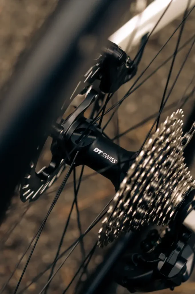 Primer plano de una rueda trasera de bicicleta que muestra el buje DT Swiss, el rotor del freno de disco y los engranajes de cassette, con parte del cuadro MMR Aelion visible al fondo, una auténtica bici asturiana.
