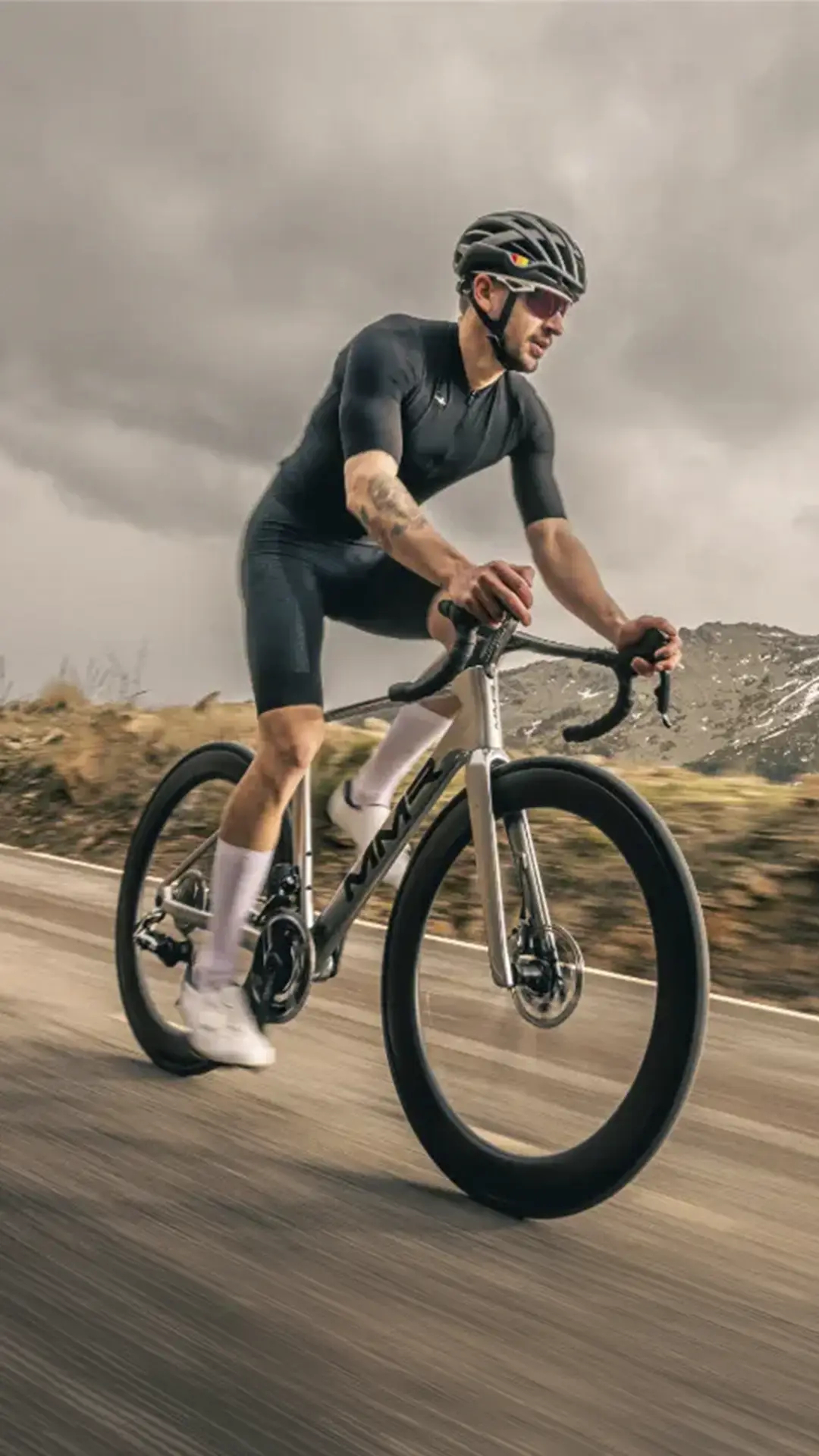 Un ciclista con ropa oscura y casco monta en una bicicleta de carretera MMR Aelion en un sendero al aire libre con montañas y cielos nublados de fondo.