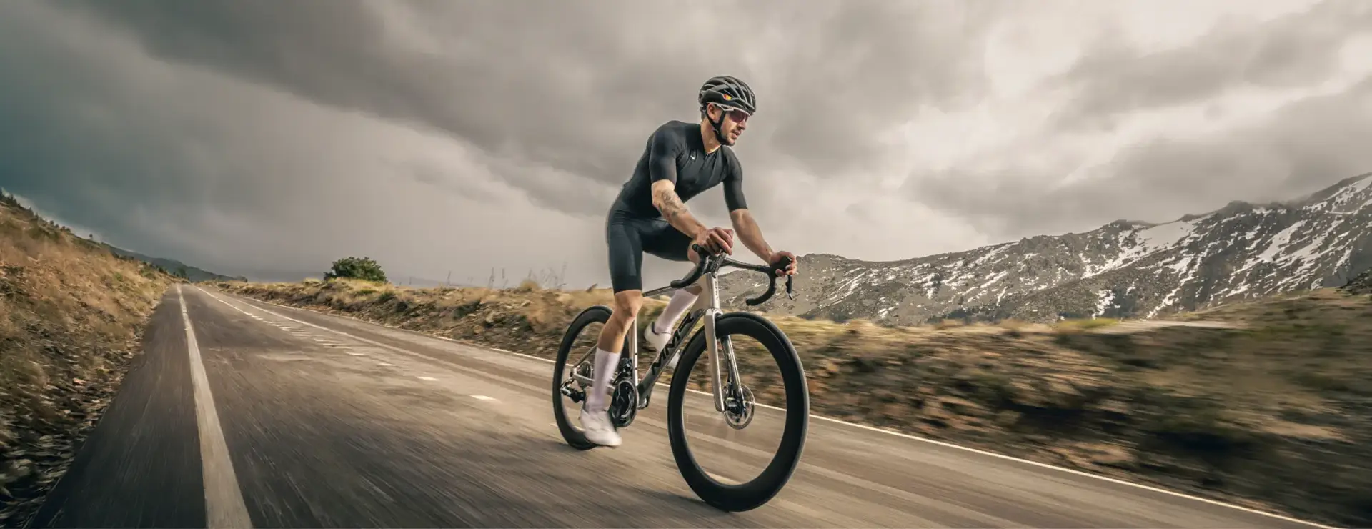Un ciclista conduce una bicicleta de carretera MMR Aelion SL por una carretera de montaña asfaltada bajo un cielo nublado, con colinas nevadas visibles al fondo.