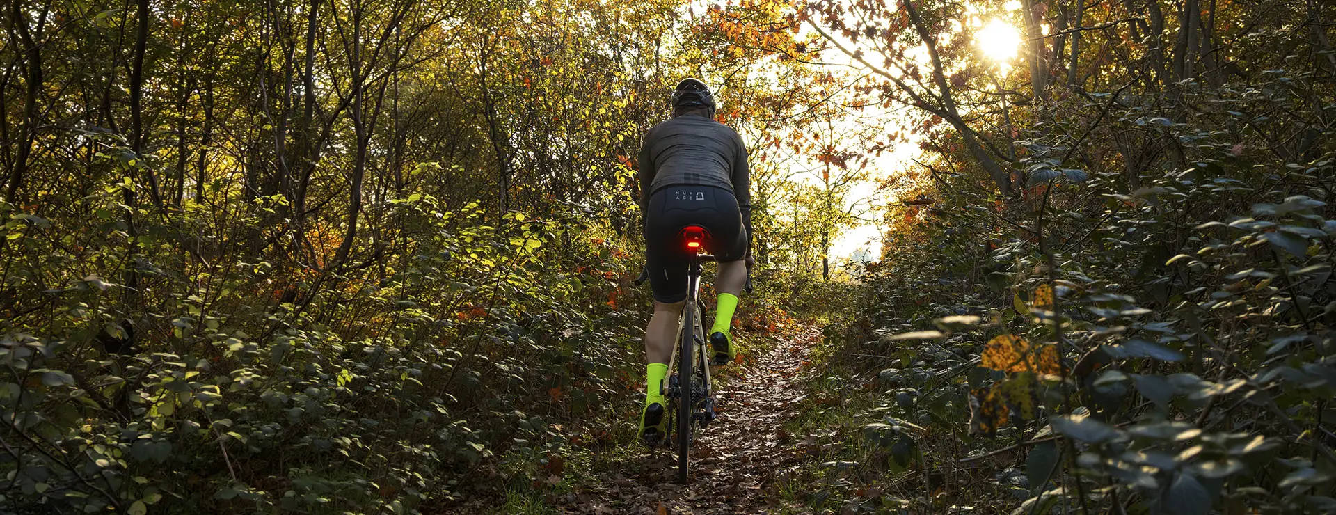 Una persona con calcetines de color amarillo brillante monta en bicicleta con un CLIP SIGMA NYTE acoplado, deslizándose por un estrecho sendero de tierra a través de una zona arbolada e iluminada por el sol.