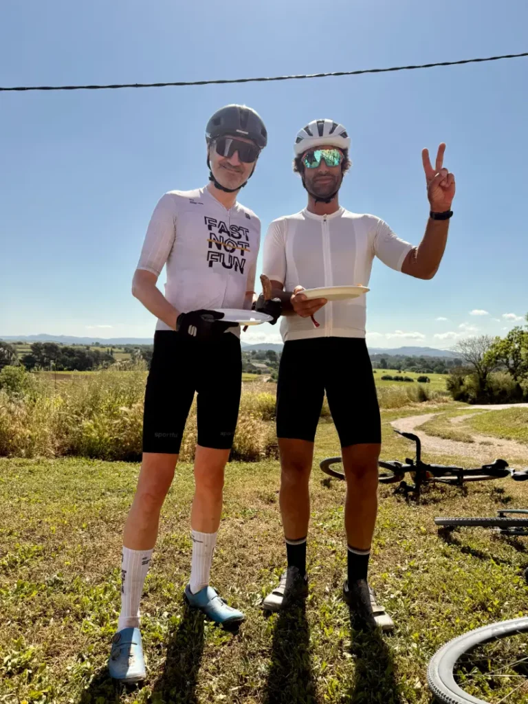 Dos ciclistas del Trek BCN Sud, con gafas de sol y casco, de pie al aire libre sobre la hierba y sosteniendo platos de comida; uno de ellos hace un gesto de paz. Al fondo se ven bicicletas y el paisaje del Penedès de Grava-2026.