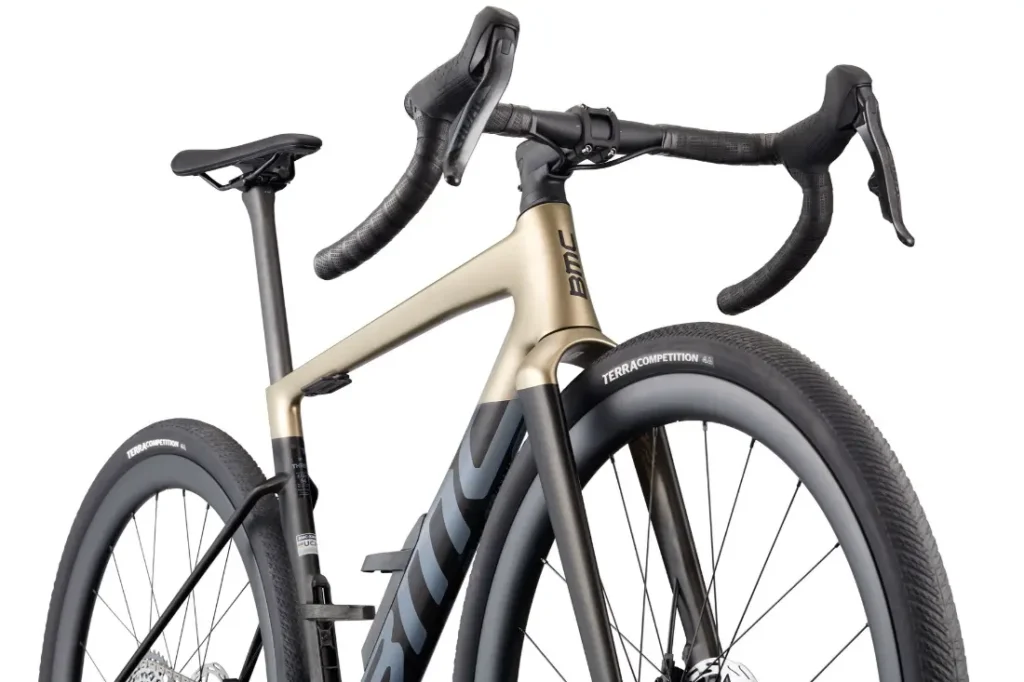 Primer plano de una BMC Kaius 01, una bicicleta rápida con manillar abatible, cuadro negro y dorado, gruesos neumáticos Terra Competition y la llamativa marca BMC.