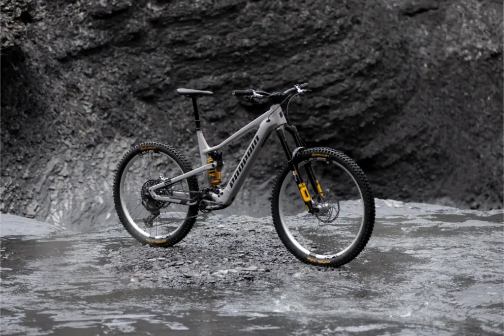 Una bicicleta de montaña Avinox de suspensión total, con cuadro gris y detalles amarillos, está aparcada sobre suelo rocoso húmedo frente a una oscura pared de roca.