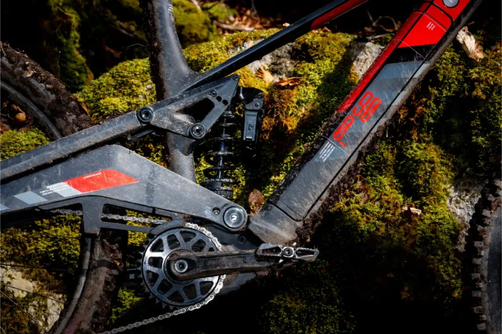 Primer plano de un cuadro de bicicleta de montaña Rotwild R.EXC que muestra la suspensión trasera, los platos y bielas -detalles perfectos para los amantes de la competición- con una roca musgosa al fondo.