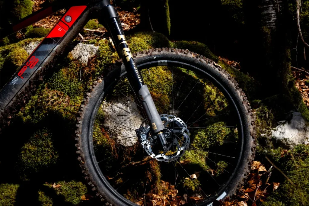 Primer plano de una rueda delantera de bicicleta de montaña Rotwild R.EXC con horquilla de suspensión FOX, lista para competir, aparcada contra rocas musgosas y árboles en un sereno entorno forestal.