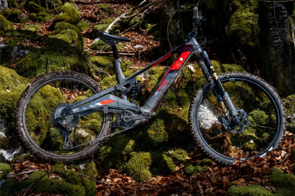 Una bicicleta de montaña Rotwild R.EXC de suspensión total con barro en los neumáticos está aparcada sobre rocas musgosas y hojas caídas en una zona boscosa, lista para competir en cualquier terreno.