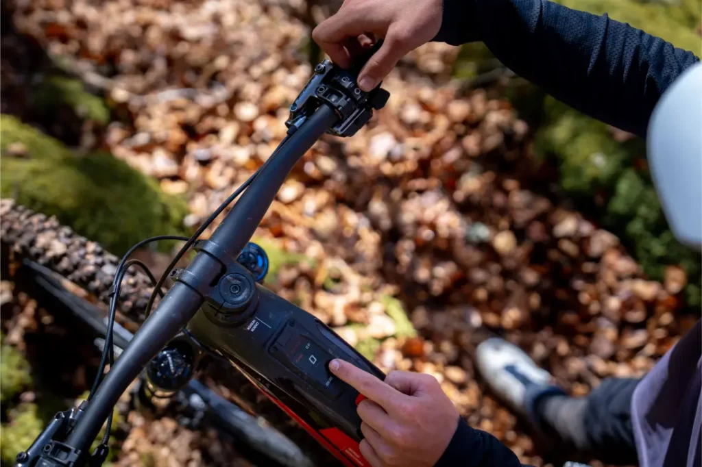 Una persona ajusta la pantalla electrónica de una bicicleta eléctrica de montaña Rotwild R.EXC mientras se encuentra en un sendero forestal cubierto de hojas, lista para competir en terrenos difíciles.