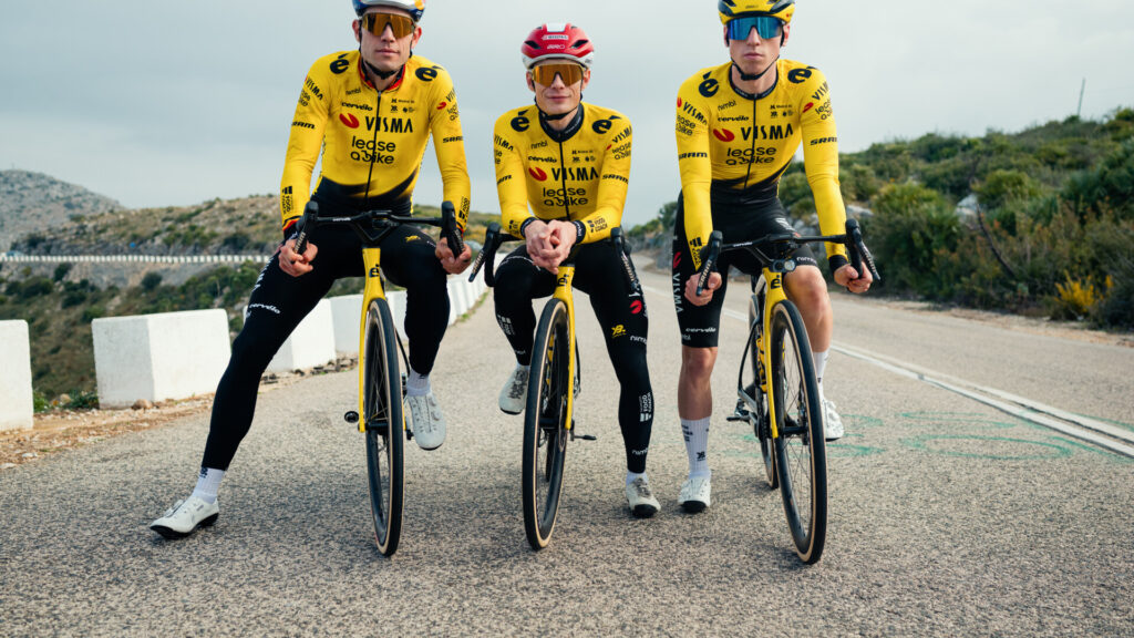 Tres ciclistas con maillots y cascos amarillos a juego del equipo Visma recorren una carretera asfaltada, utilizando un sensor de entrenamiento para optimizar su entrenamiento ciclista con un impresionante telón de fondo montañoso.