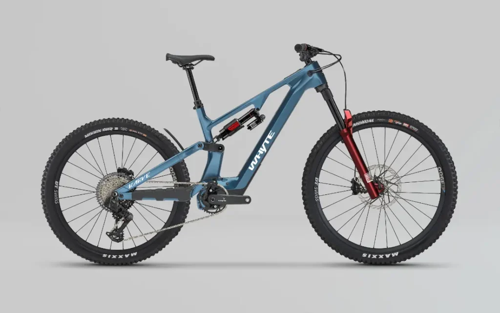 Una bicicleta de montaña Avinox azul con suspensión total y detalles en negro y rojo, con frenos de disco, neumáticos de tacos y un tubo superior inclinado, sobre un fondo gris liso.