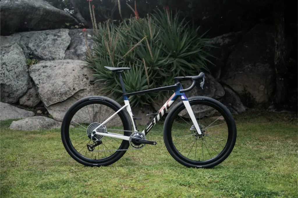 Una BMC Kaius 01 bicicleta rápida negra, blanca y azul está aparcada sobre hierba delante de rocas y plantas verdes al aire libre.