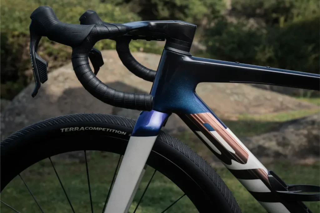 Primer plano de una BMC Kaius 01, una bicicleta rápida, destacando el manillar, la rueda delantera y parte del cuadro, en exteriores con rocas y vegetación de fondo.