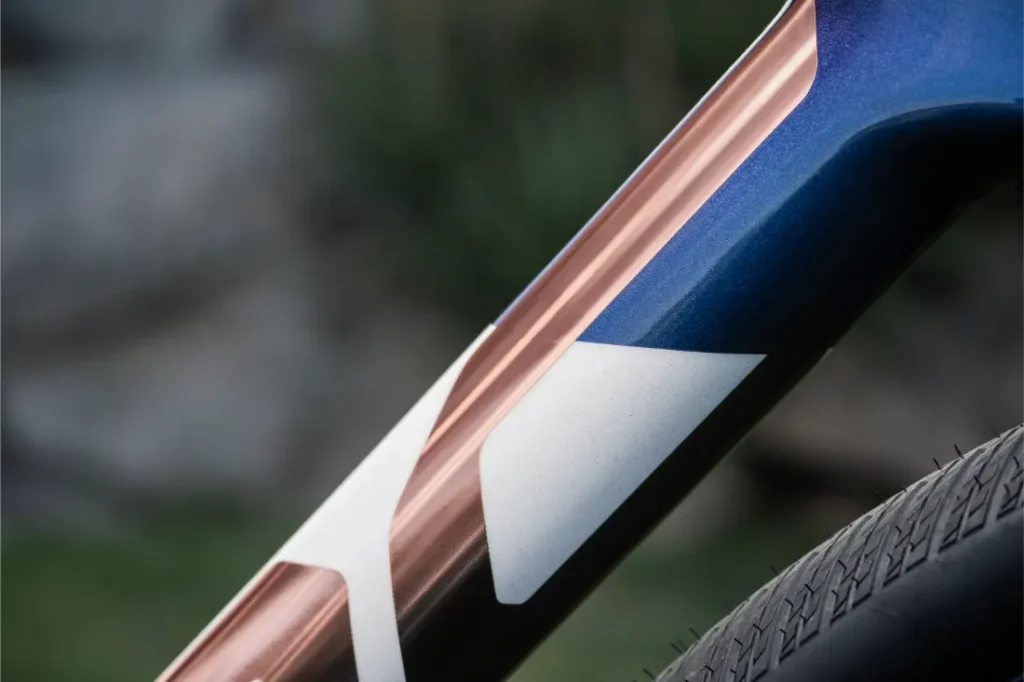 Primer plano del cuadro de la bicicleta BMC Kaius 01 con una combinación de colores cobre metálico y azul con un diseño geométrico blanco, junto a un neumático de bicicleta texturizado, perfecto para cualquiera que busque una bicicleta rápida y actual.