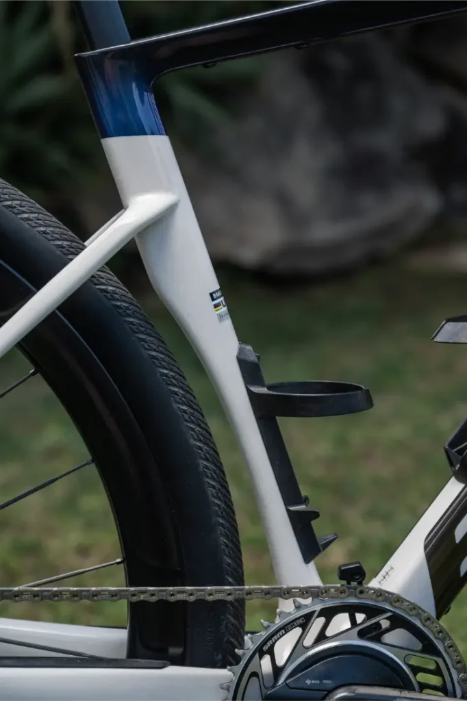 Primer plano del cuadro de una BMC Kaius 01 bicicleta rápida, mostrando el tubo del sillín, la rueda trasera, el portabidón y parte de la cadena y el plato, con hierba y una gran roca al fondo.
