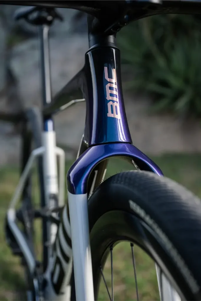 Primer plano de una bicicleta de carretera BMC Kaius 01 que muestra la horquilla delantera, el neumático y parte del cuadro, con vegetación exterior en el fondo difuminado: una bicicleta rápida ideal para cualquier paseo.