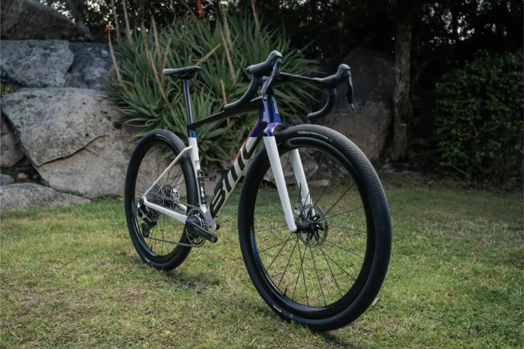 La actual bicicleta BMC Kaius 01, con guía de descenso y neumáticos largos, está estacionada en la parrilla junto a grandes rocas y muy verde.