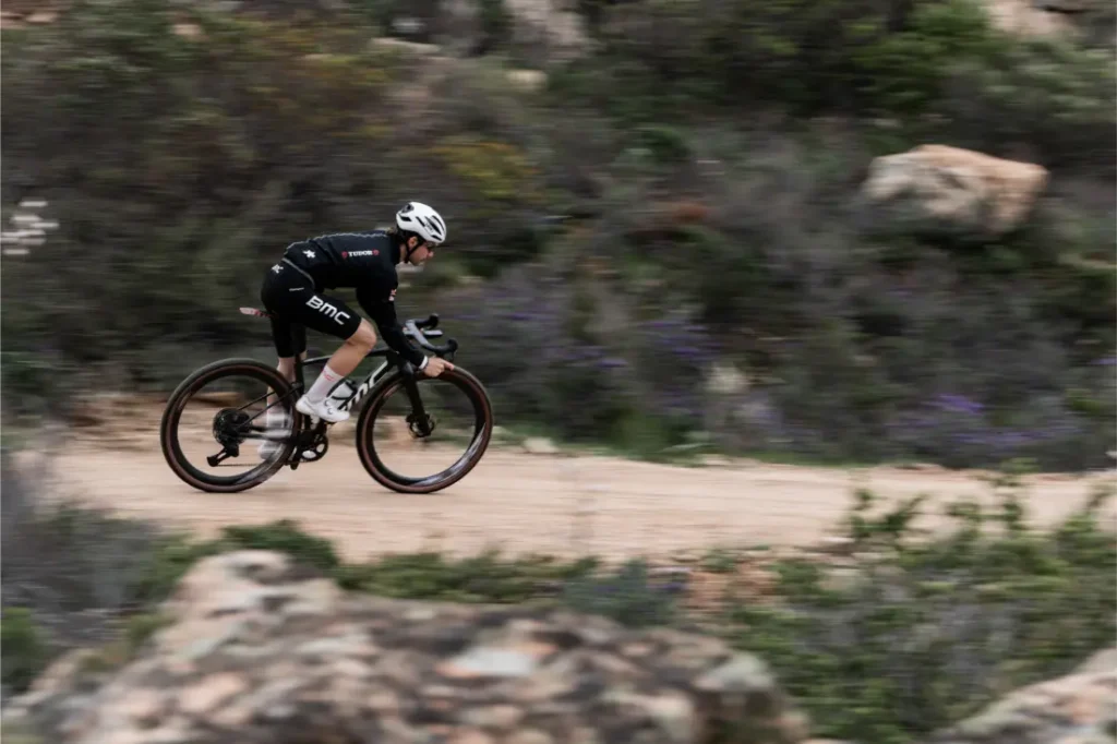 Un ciclista con la equipación del equipo BMC conduce una bicicleta rápida, la BMC Kaius 01, por un camino de tierra a través de un paisaje seco y rocoso con un follaje borroso.