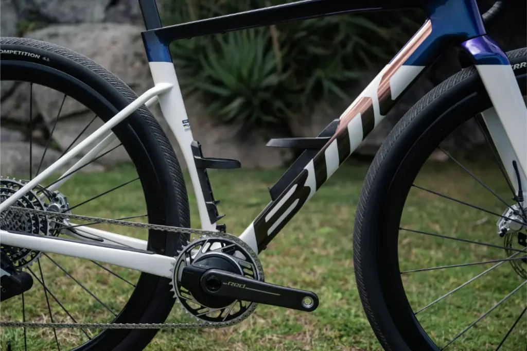 Primer plano de la BMC Kaius 01, una bicicleta rápida, destacando su cuadro, transmisión y frenos de disco, con hierba y plantas de fondo.