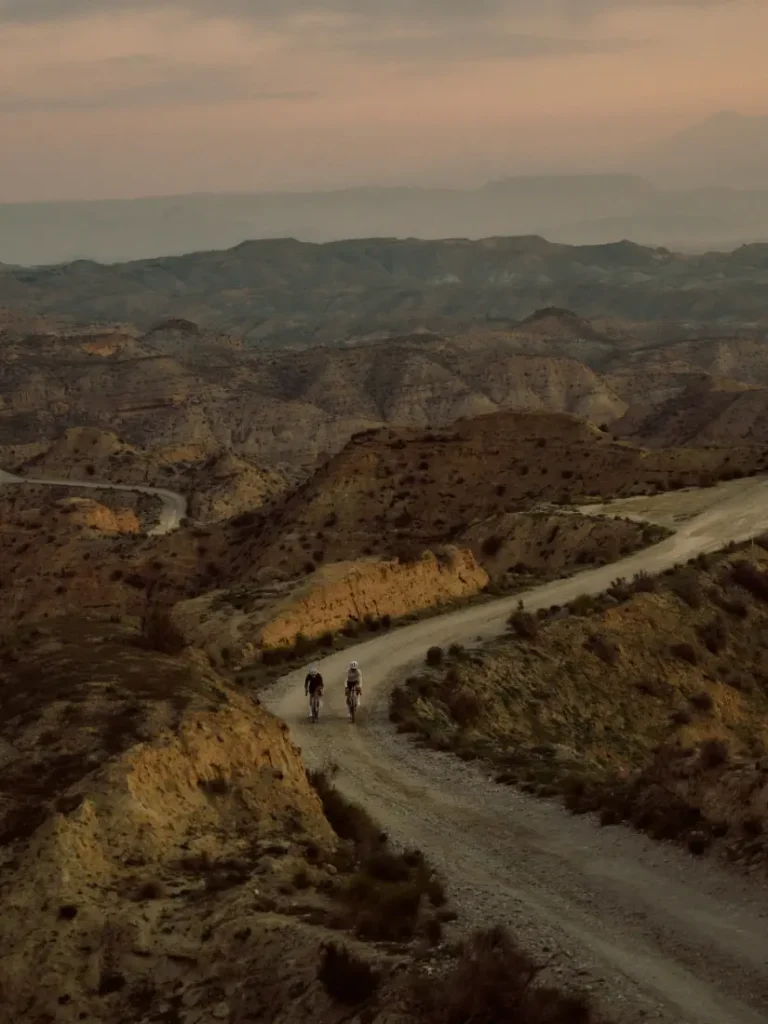 Dos ciclistas recorren un sinuoso camino de tierra a través de un paisaje accidentado y montañoso al anochecer; uno de ellos va en una BMC Kaius 01, disfrutando de la emoción de esta bicicleta rápida bajo un cielo nublado.