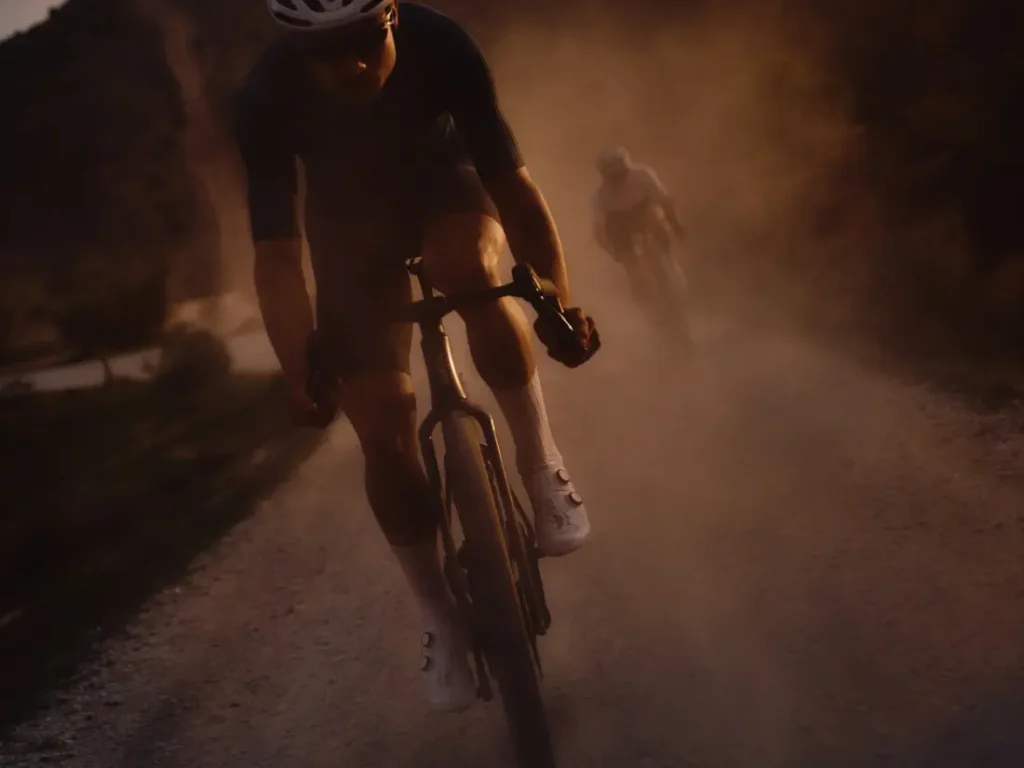 Dos ciclistas circulan por una polvorienta carretera de grava al atardecer. El ciclista que va en cabeza, en primer plano, avanza a toda velocidad en una BMC Kaius 01 bicicleta rápida, mientras otro ciclista le sigue de cerca.