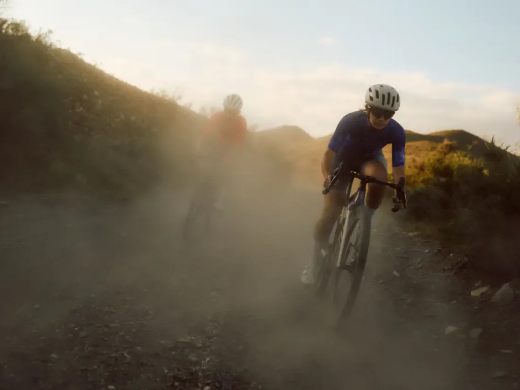 Dos ciclistas montan en bicicletas BMC Kaius 01 por un sendero de tierra, levantando polvo mientras atraviesan a toda velocidad un paisaje de colinas bajo un cielo parcialmente nublado.