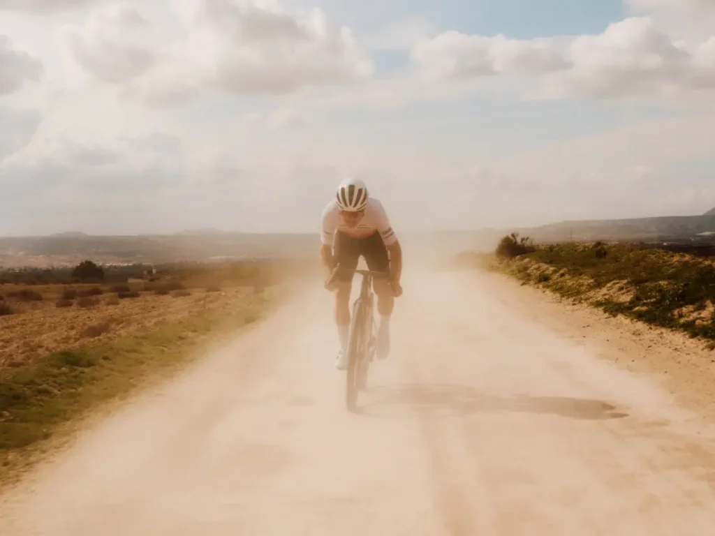 Un ciclista con casco y gafas de sol conduce una bicicleta rápida, la BMC Kaius 01, por un polvoriento camino rural de tierra bajo un cielo parcialmente nublado.