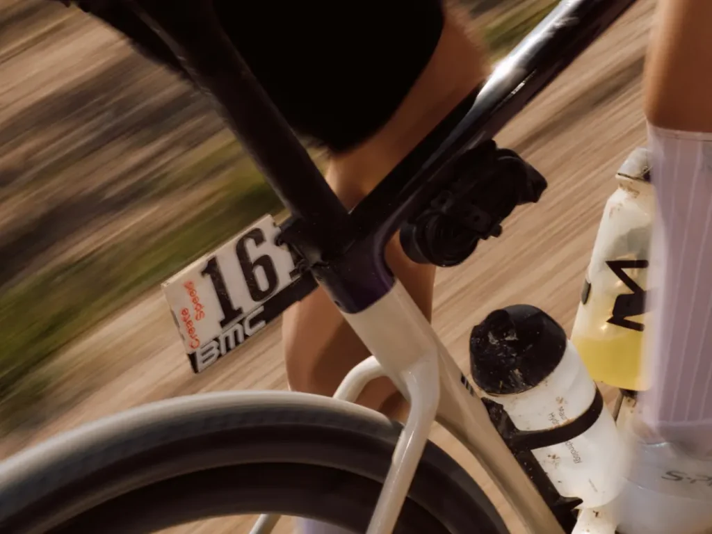 Primer plano de una persona montada en una BMC Kaius 01 blanca, dorsal 163, botellas de agua colocadas y fondo borroso que indica el movimiento de esta bicicleta rápida.