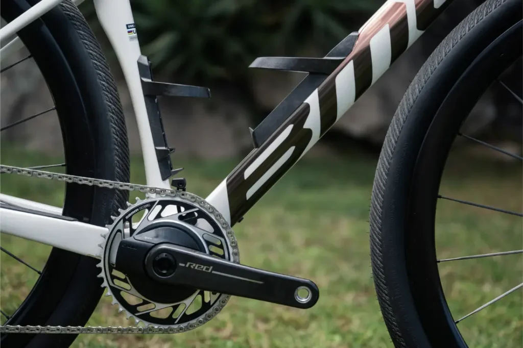 Primer plano de los platos, la cadena y el cuadro de una BMC Kaius 01, con la bicicleta rápida sobre hierba y follaje difuminado al fondo.