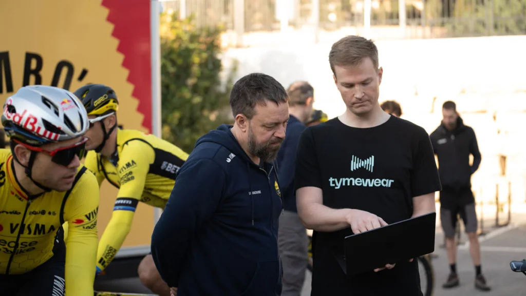 Varios hombres permanecen al aire libre; uno de ellos, con una camiseta Tyme Wear negra, muestra la pantalla de un ordenador portátil a otro hombre, haciendo una demostración de un sensor de entrenamiento, mientras unos ciclistas vestidos de amarillo esperan cerca.