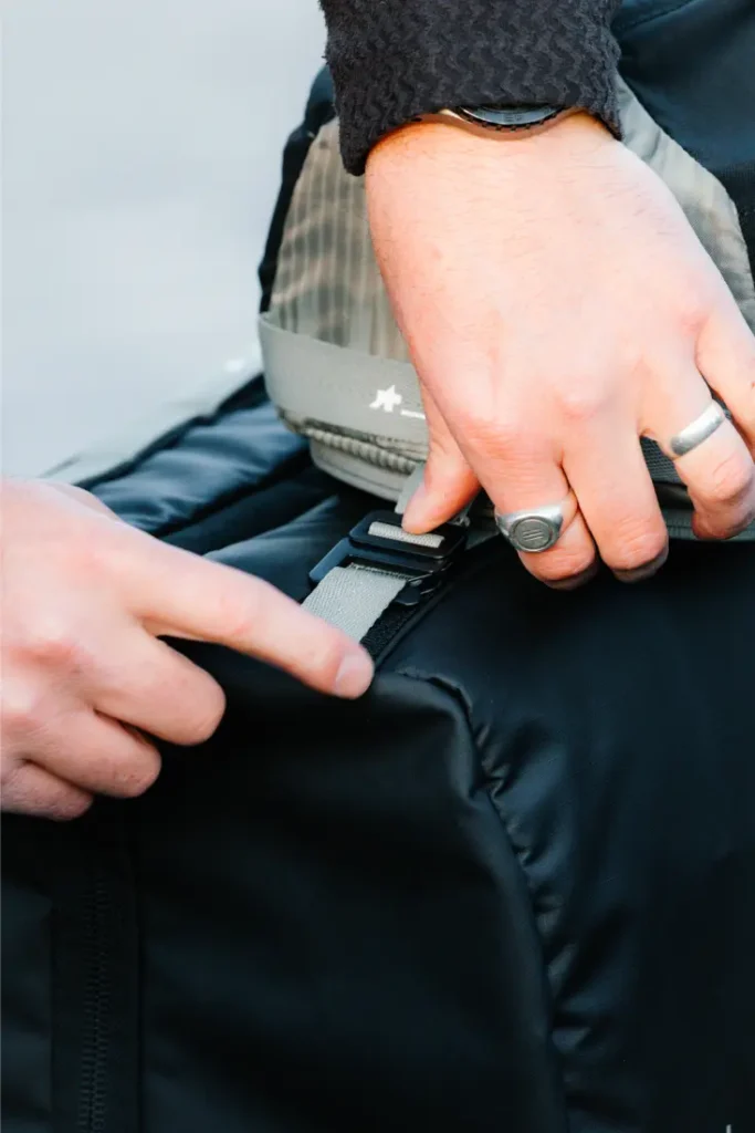 Primer plano de dos manos ajustando una hebilla en un bolso negro ASSOS x Db -perfecto para ciclistas- una persona lleva anillos y un reloj, destacando el elegante sistema de viaje.