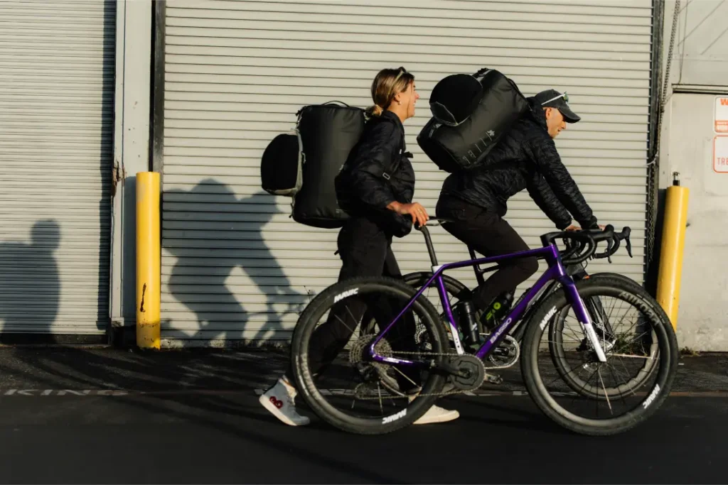 Dos ciclistas con mochilas caminan y montan en bicicleta junto a la puerta de un garaje de chapa ondulada, proyectando largas sombras en el suelo: una escena inspirada en el sistema de viaje ASSOS x Db.