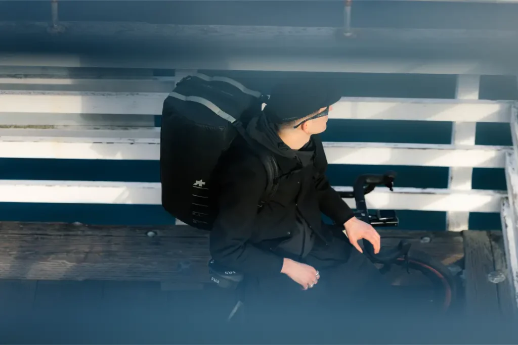 Una persona vestida de negro, con una mochila ASSOS x Db, se sienta en una bicicleta cerca de una barandilla blanca, vista desde arriba a través de unas barras horizontales difuminadas, que captan la esencia de los ciclistas en movimiento y de un innovador sistema de viaje.
