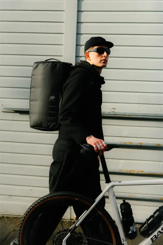 Una persona vestida con ropa negra, gorra y gafas de sol se sitúa junto a una bicicleta con una gran mochila negra ASSOS x Db, encarnando el espíritu del sistema de viaje para ciclistas contra una pared metálica de color claro.