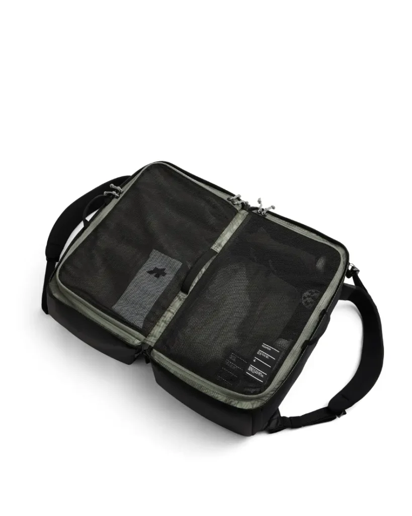 Una bolsa de viaje ASSOS x Db negra abierta en plano, mostrando dos compartimentos de malla con cremallera con ropa en su interior, perfecta para ciclistas y un organizado sistema de viaje, colocada sobre un fondo blanco.