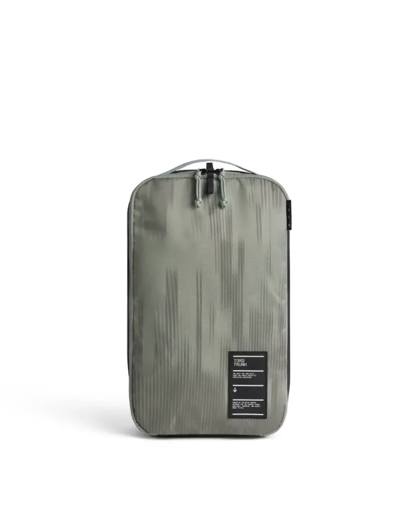 Cubo de embalaje rectangular verde oliva de Db con sutiles dibujos verticales, cierre de cremallera en la parte superior y una pequeña etiqueta negra con texto blanco en la esquina inferior derecha: un complemento ideal para cualquier sistema de viaje.