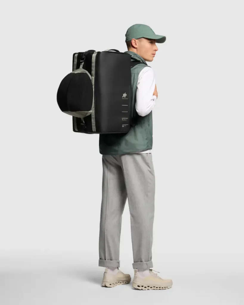 Persona con gorra verde, chaleco verde, camisa blanca, pantalón gris y zapatos beige, que lleva una mochila negra rectangular grande ASSOS x Db, parte de un innovador sistema de viaje ideal para ciclistas.