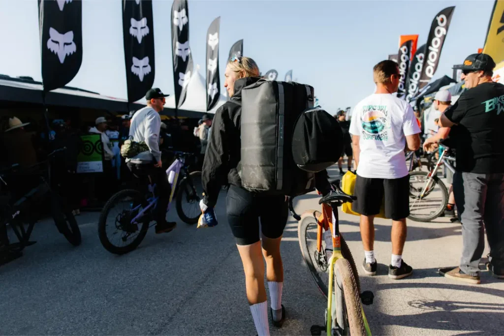 Una persona con una gran mochila ASSOS x Db y una bicicleta camina a través de un concurrido evento al aire libre lleno de ciclistas, bicicletas, banderas y gente con atuendo informal.