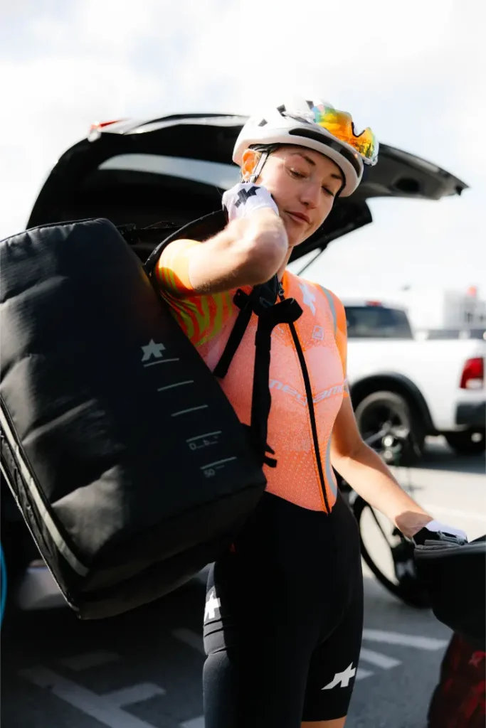 Un ciclista con casco y gafas de sol, luciendo el sistema de viaje ASSOS x Db, lleva una gran bolsa negra sobre un hombro en un aparcamiento con el maletero de un coche abierto detrás.