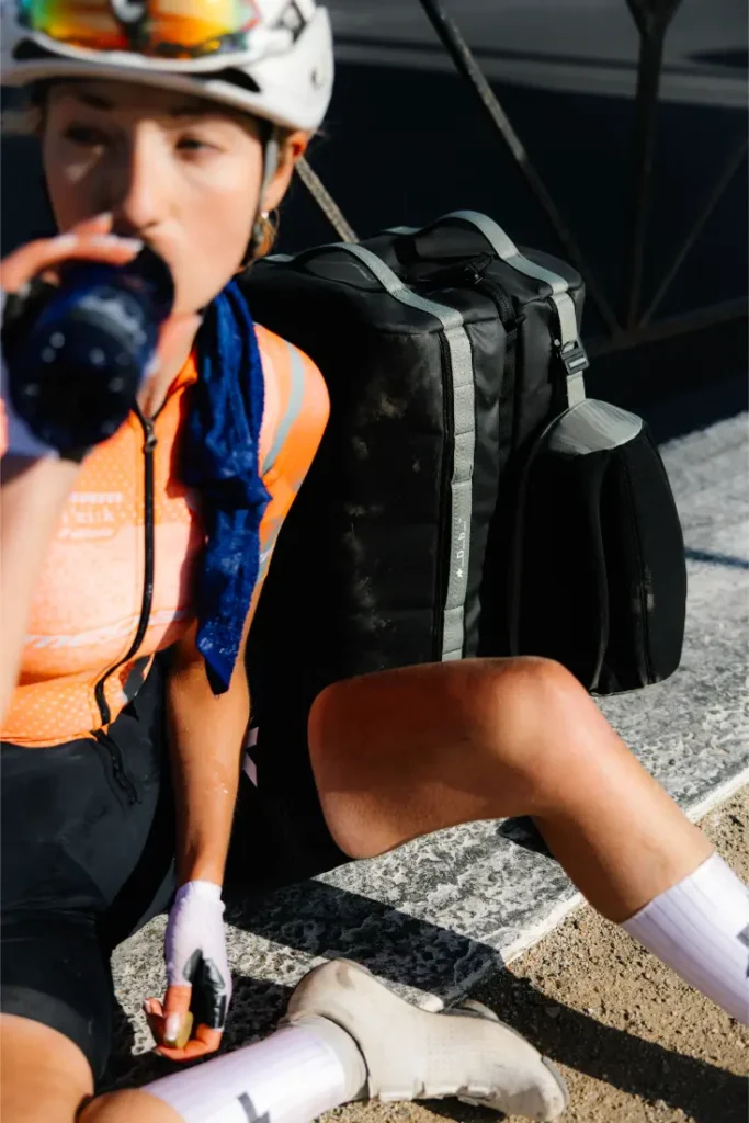 Un ciclista con casco y maillot naranja sentado en un bordillo, bebiendo de una botella, con una bolsa de deporte ASSOS x Db sistema de viaje a su lado.