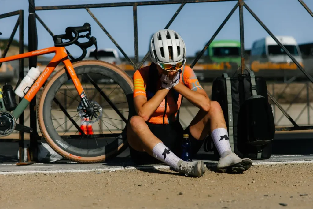 Ciclista con maillot naranja y casco sentado en el suelo junto a una bicicleta, apoyado contra una valla con la cabeza entre las manos: bolsas del sistema de viaje ASSOS x Db y botellas de agua cerca, captando la fatiga que muchos ciclistas conocen.