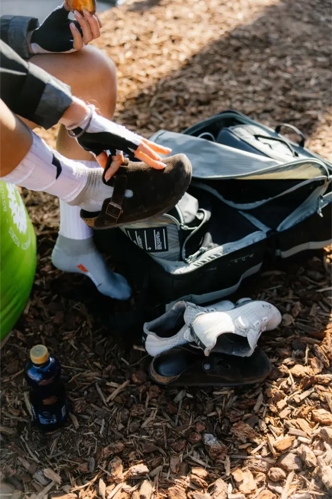 Una persona con guantes y calcetines blancos se calza unas zapatillas de escalada junto a una bolsa abierta del sistema de viaje ASSOS x Db, con otro par de zapatillas y una botella en el suelo: una organización perfecta para ciclistas.