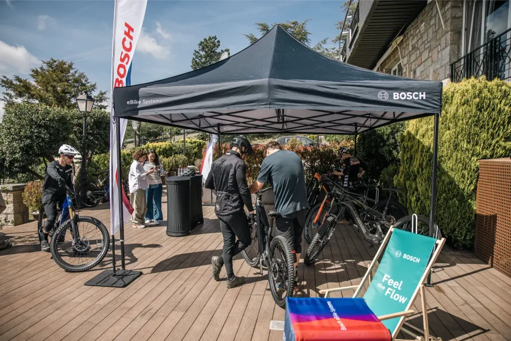 La gente se reúne bajo una carpa con la marca Bosch en la que se exponen bicicletas eléctricas sobre una cubierta de madera, explorando la experiencia sobre la bici con el Bosch Performance Upgrade para más potencia. Cerca hay una tumbona y material promocional.