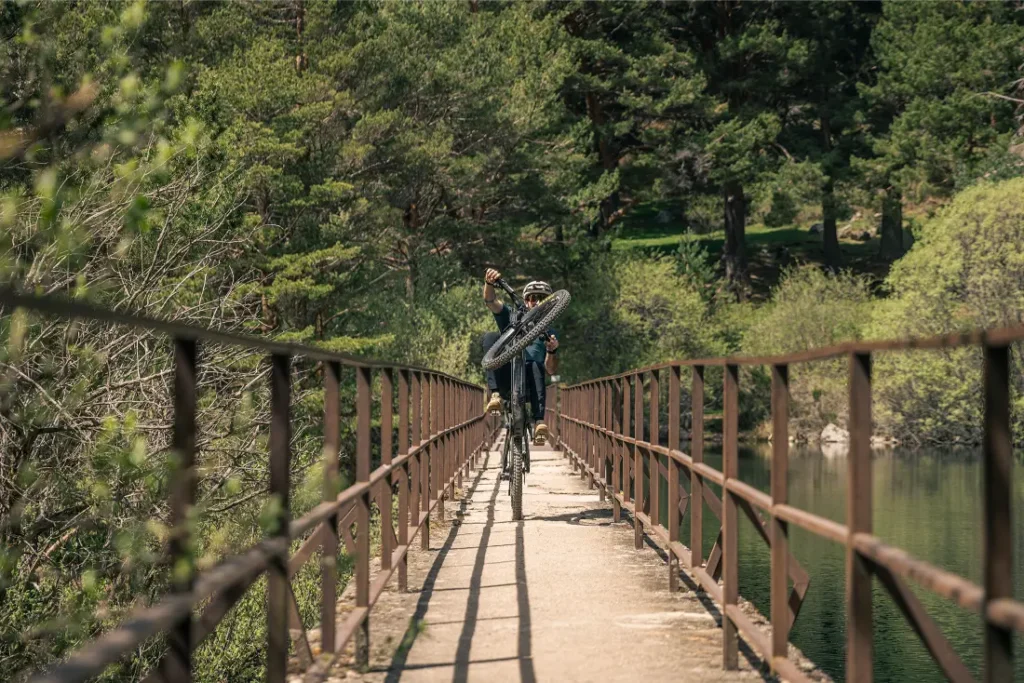 Dos personas cargadas con bicicletas de montaña atraviesan un puente estrecho rodeado de árboles y vegetación en un día soleado, dispuestas a vivir una experiencia sobre la bici con más potencia gracias a la Bosch Performance Upgrade.
